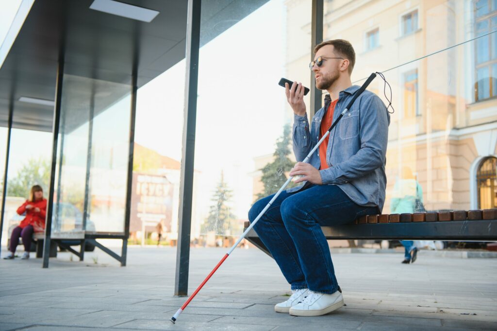 Young blind man with smartphone sitting on bench in park in city, calling.