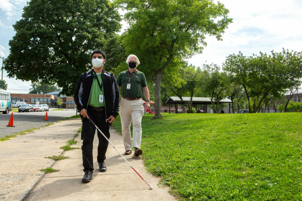 A visually impaired man using a white cane with a sighted guide behind him.