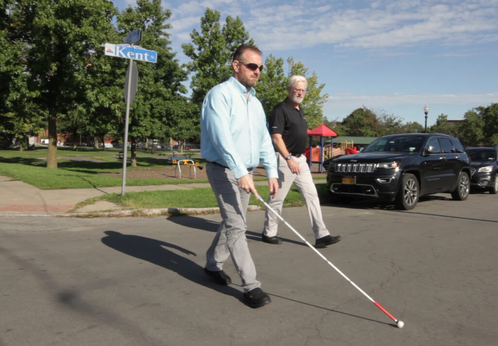 A visually impaired man using a white cane to cross the street. A sighted guide is walking next to the visually impaired man.